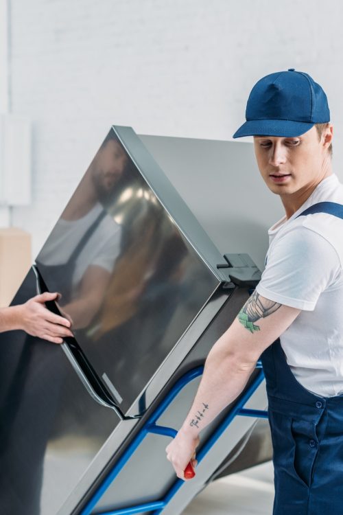 two-movers-in-uniform-using-hand-truck-while-transporting-refrigerator-in-apartment